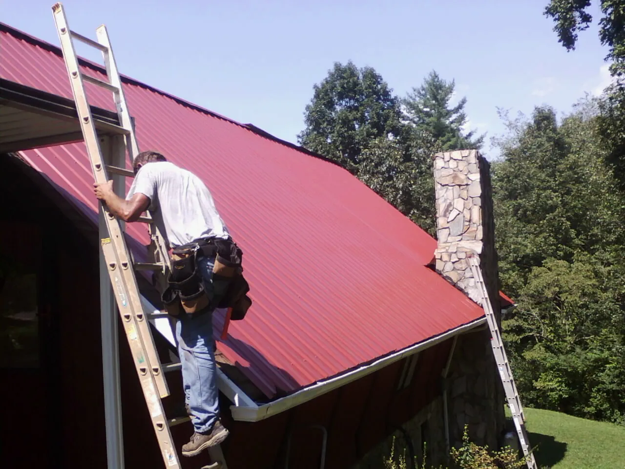 Roofer on a ladder beside a red metal roof and stone chimney — Soffit & Fascia Repair by Roofing Brothers in Vina